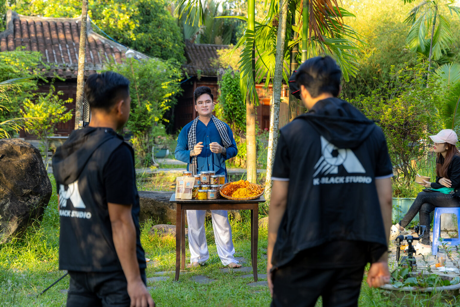 Hai thành viên ekip K-BLACK quay nam MC áo xanh chàm giới thiệu món ăn ngoài sân vườn