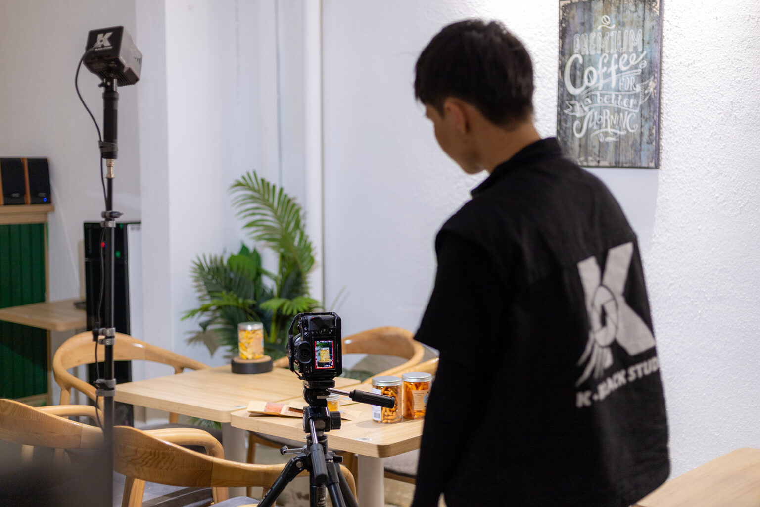 Nhân viên K-BLACK Studio setup máy quay trên tripod cạnh đèn LED để quay sản phẩm trong quán cà phê