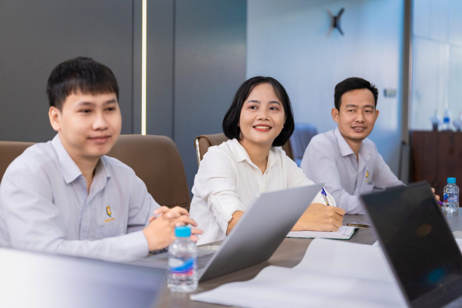 Ảnh profile công ty chuyên nghiệp: ba nhân viên mặc sơ mi đồng phục ngồi bàn họp với laptop, mỉm cười tự nhiên