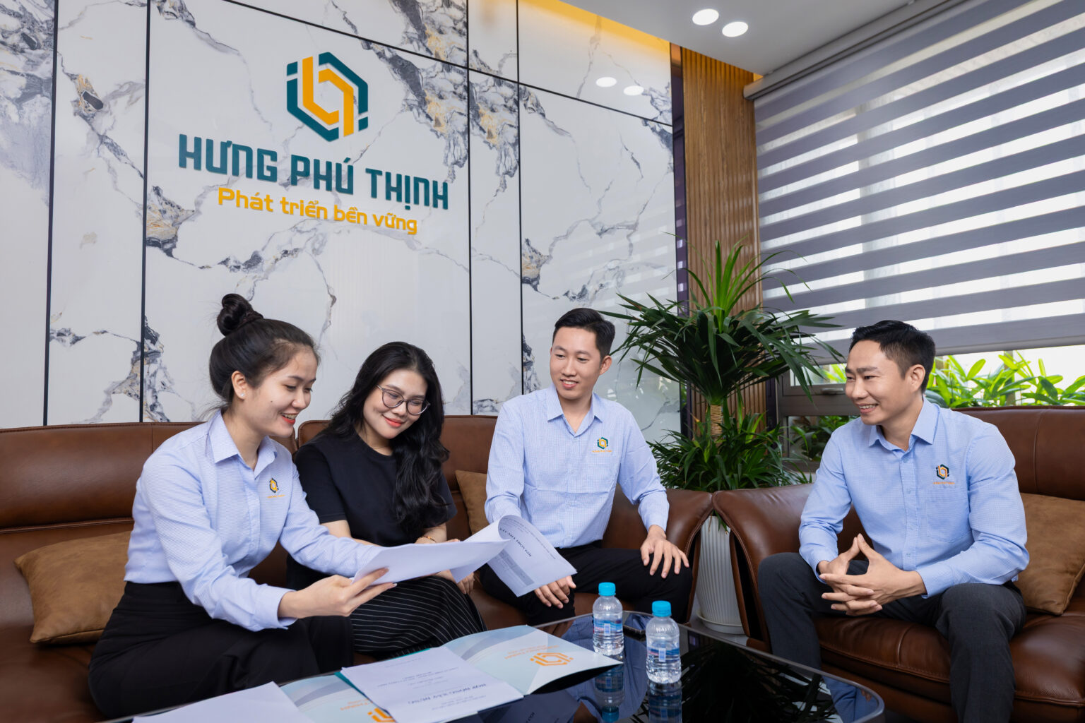 Ảnh profile công ty chuyên nghiệp: nhân sự đồng phục xanh nhạt ngồi sofa da xem tài liệu trước tường đá cẩm thạch có logo công ty