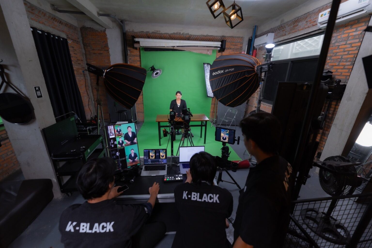 Toàn cảnh livestream sự kiện online tại K-BLACK Studio: nữ host ngồi bàn trước phông xanh, hai softbox Godox, kỹ thuật viên áo đen ở tiền cảnh