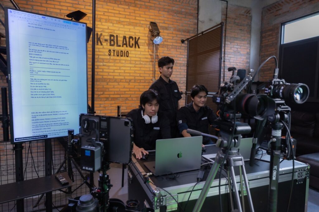 Ekip K-BLACK áo đen chuẩn bị livestream sự kiện online tại studio tường gạch, một người đeo tai nghe ngồi trước MacBook, màn hình lớn hiển thị kịch bản